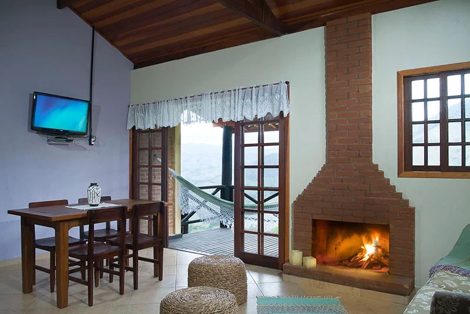 Sala com lareira acesa e porta aberta para a varanda com rede e vista da serra