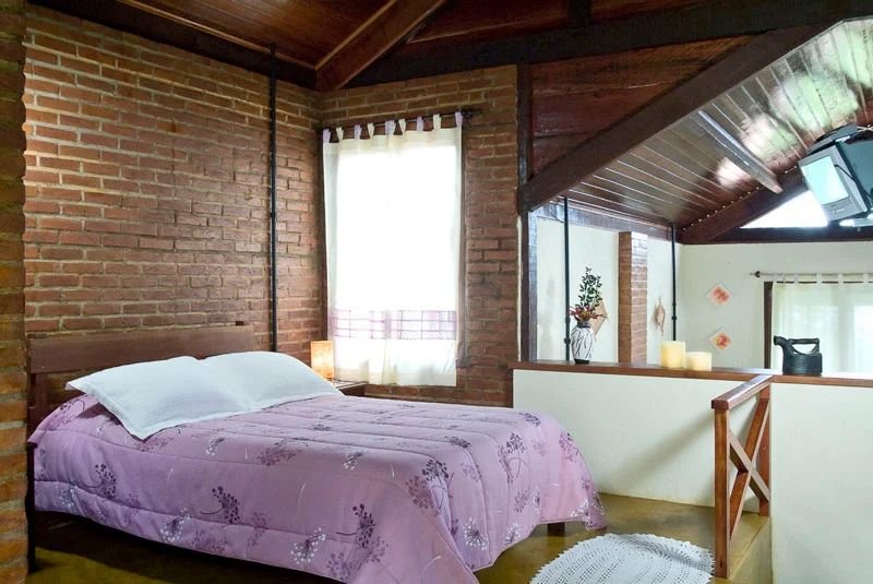 Cama de casal no mezanino com parede de tijolos e janela de cortina clara